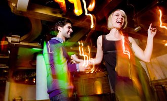 A couple is dancing happily in a lively environment with colorful lights. The atmosphere is energetic and inviting. | © Aparthotel Kleinwalsertal Haller GmbH & Co KG
