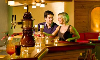 A cozy bar area with a couple conversing. In the background, various drinks and warm wooden decor can be seen. | © Aparthotel Kleinwalsertal Haller GmbH & Co KG