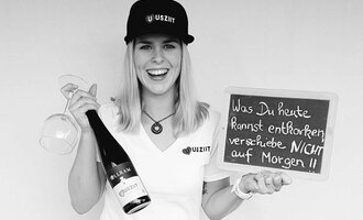 A cheerful woman is holding a glass and a bottle of champagne in her hand. She is standing next to a board with the inscription: "What you can remove today, do NOT postpone until tomorrow!!". | © Uusziit | Chantal Kessler