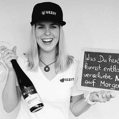 A cheerful woman is holding a glass and a bottle of champagne in her hand. She is standing next to a board with the inscription: "What you can remove today, do NOT postpone until tomorrow!!". | © Uusziit | Chantal Kessler