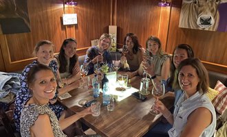 A group of eight women is sitting at a table and raising glasses. It is a cheerful atmosphere, surrounded by wooden decor and soft light. | © Uusziit | Chantal Kessler