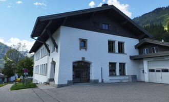 Ein modernes, weißes Gebäude mit einem großen Dach. Es befindet sich in einer bergigen Umgebung mit grünem Gras und Bäumen. | © Kleinwalsertal Tourismus | N. Lughammer