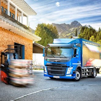 A modern truck stands in front of a wooden building with mountains in the background. A forklift is moving nearby while the sun is shining. | © NUFA | Stefan Kothner