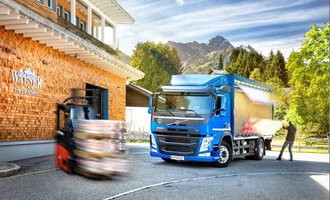 A modern truck stands in front of a wooden building with mountains in the background. A forklift is moving nearby while the sun is shining. | © NUFA | Stefan Kothner