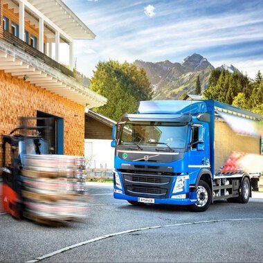 A modern truck stands in front of a wooden building with mountains in the background. A forklift is moving nearby while the sun is shining. | © NUFA | Stefan Kothner