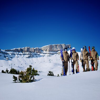 A group of skiers stands on a snow-covered area. In the background, you can see impressive mountain peaks and a clear blue sky. | © Walser Skischule | Herbert Jochum
