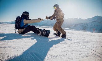 Two snowboarders on a snowy slope. One is helping the other, who is sitting on the ground. | © Walser Skischule | Ole Zumpolle