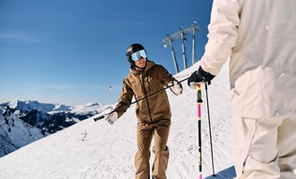 A skier in a brown ski jacket is standing on a snow-covered slope. In the background, there are snow-covered mountains and a blue sky. | © Walser Skischule | Oliver Farys