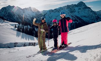 Three skiers are standing on a snowy slope and waving at the camera. In the background, impressive mountains and a clear sky can be seen. | © Walser Skischule | Ole Zumpolle
