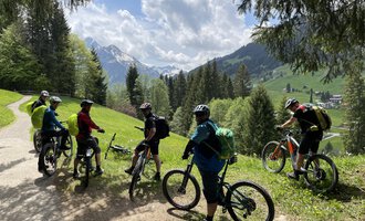 A group of cyclists is standing on a path in the mountains. The surroundings are green with forest and an impressive mountain landscape in the background. | © WalserBike Tours | Christian Gutermuth