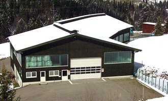 A large, modern building with a black facade and a snow-covered roof. Surrounded by snowy trees and a peaceful landscape. | © Gemeinde Mittelberg