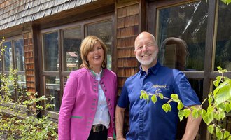 The image shows two people standing in front of a wooden house. The woman is wearing a pink jacket, the man a blue shirt, both smiling kindly. | © Wirtshaus Hoheneck | Klaus Ruland