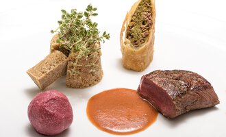 An artistically arranged plate with juicy meat, a green herb decoration, and various side dishes. The dishes are appealingly combined and have vibrant colors. | © Wirtshaus Hoheneck | Klaus Ruland