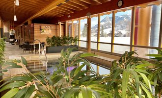 An elegant wellness area with a small pool and plants. Large windows offer a view of the surrounding mountains. | © WONNEMAR Sonthofen | Julia Sondergelt