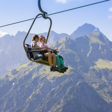 Two people are sitting in a gondola on their way over the mountains. The landscape in the background is majestic with green meadows and high peaks. | © Kleinwalsertal Tourismus | Frank Drechsel